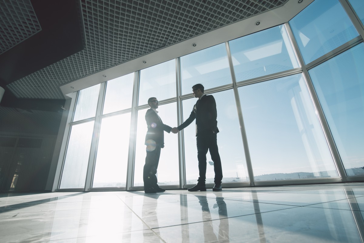 Two men shake hands in front of large windows with sun shining through132400690