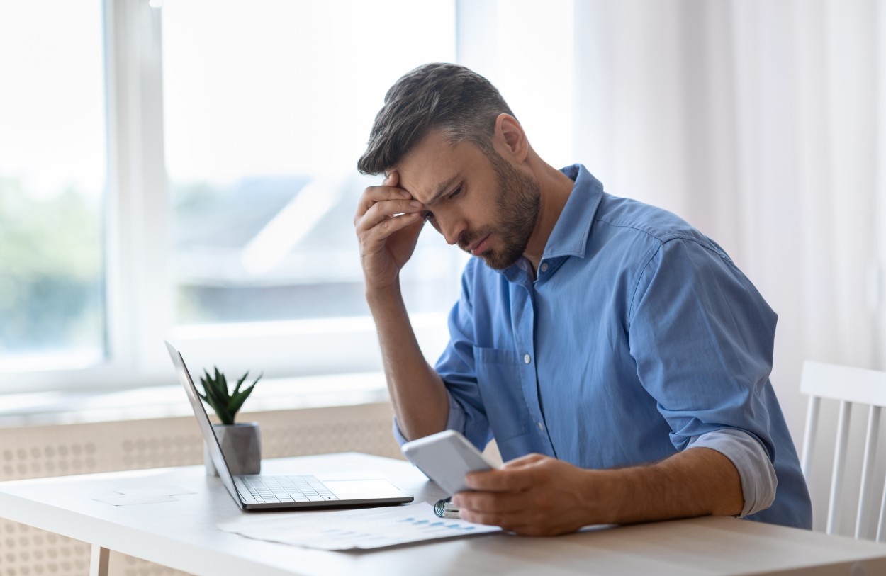 concerned man looks at laptop and phone 395360650